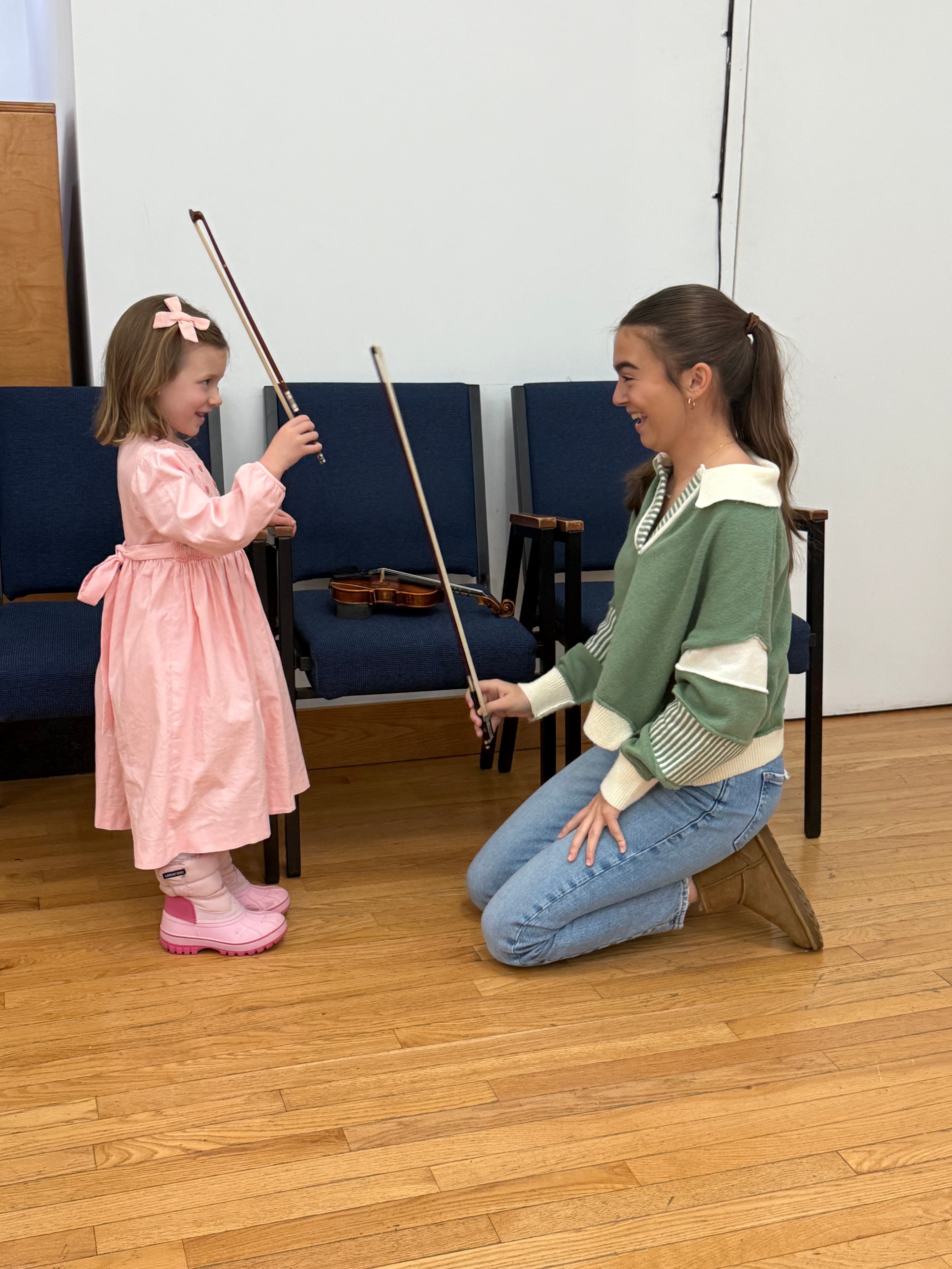 A teacher kneeling to guide a young student through her first bow strokes